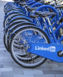 A row of blue bicycles is aligned on a tiled surface, each bicycle featuring the LinkedIn logo prominently displayed on the side panels. The bicycles are neatly arranged, suggesting they are part of a bike-sharing program or rental service.