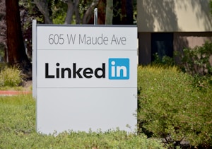 A sign with the address 605 W Maude Ave and the LinkedIn logo is surrounded by greenery and trees.