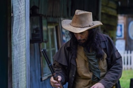 A person with long hair is wearing a cowboy hat and traditional Western attire. They are holding a double-barreled shotgun while standing outdoors near a rustic wooden building. The setting appears to evoke an Old West theme with wooden textures and ambient lighting.