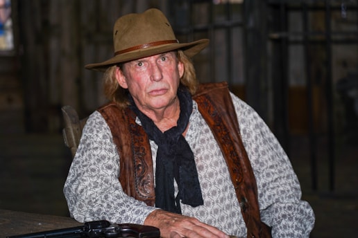An older man with a rugged appearance is dressed in Western attire, including a brown hat, patterned shirt, leather vest, and black scarf. He is seated indoors, possibly in a dimly lit, rustic setting. A revolver is partially visible on the table in front of him, enhancing the cowboy theme.