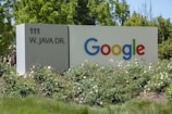 A large sign with 'Google' written in colorful letters is positioned next to '111 W. Java Dr.' The sign is surrounded by green foliage and white flowers, set against a backdrop of trees under clear skies.