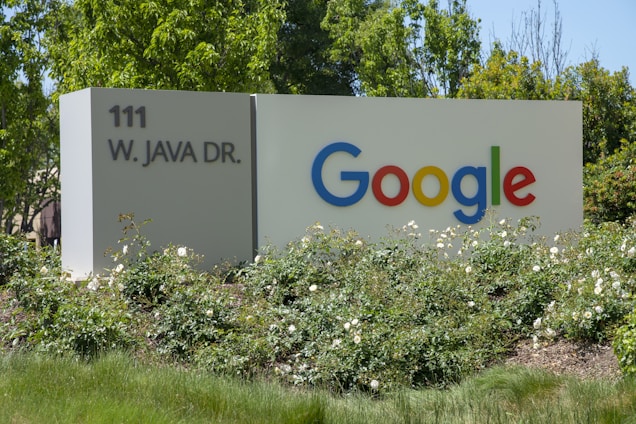 A large sign with 'Google' written in colorful letters is positioned next to '111 W. Java Dr.' The sign is surrounded by green foliage and white flowers, set against a backdrop of trees under clear skies.