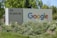 A large sign with 'Google' written in colorful letters is positioned next to '111 W. Java Dr.' The sign is surrounded by green foliage and white flowers, set against a backdrop of trees under clear skies.