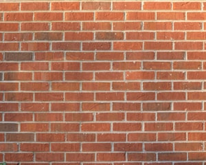 A brick wall consisting of uniform red bricks with mortar lines forming a grid pattern. The surface appears slightly weathered with minor cracks and discoloration in a few areas.