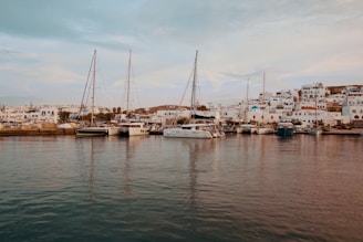 Sailboats are docked at a marina with a charming village in the background. The village features white buildings with blue accents, typical of Mediterranean architecture, set against a gently sloping hillside. The calm water of the marina reflects the boats and buildings, creating a serene and picturesque scene.