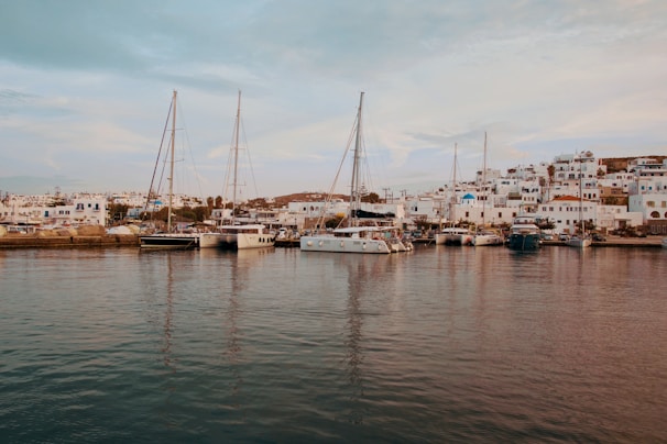 Sailboats are docked at a marina with a charming village in the background. The village features white buildings with blue accents, typical of Mediterranean architecture, set against a gently sloping hillside. The calm water of the marina reflects the boats and buildings, creating a serene and picturesque scene.