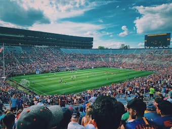 A large stadium filled with an enthusiastic crowd watching a soccer match. The lush green field is surrounded by full stands, with spectators keenly observing the players in action. The sky is partly cloudy with a vibrant blue hue, and there is a big screen displaying information on one end of the stadium. The general ambiance is lively and engaging.