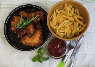 A black skillet contains four pieces of roasted chicken adorned with a sprig of fresh rosemary, while a brown bowl holds a generous serving of golden french fries. A small glass dish filled with ketchup is positioned beside silverware, all set on a green-striped fabric.