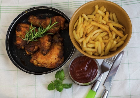 A black skillet contains four pieces of roasted chicken adorned with a sprig of fresh rosemary, while a brown bowl holds a generous serving of golden french fries. A small glass dish filled with ketchup is positioned beside silverware, all set on a green-striped fabric.