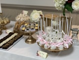 An elegantly arranged dessert table featuring a variety of sweets on golden stands. White cake pops decorated with pink ribbons are prominently displayed. Surrounding them are Ferrero Rocher chocolates in delicate wrappers and an assortment of pastries, including chocolate brownies with toppings. In the background, large white and red roses complement the table's aesthetic.