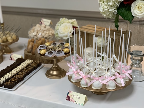 An elegantly arranged dessert table featuring a variety of sweets on golden stands. White cake pops decorated with pink ribbons are prominently displayed. Surrounding them are Ferrero Rocher chocolates in delicate wrappers and an assortment of pastries, including chocolate brownies with toppings. In the background, large white and red roses complement the table's aesthetic.
