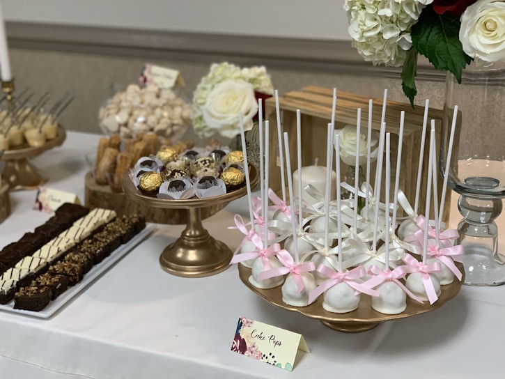An elegantly arranged dessert table featuring a variety of sweets on golden stands. White cake pops decorated with pink ribbons are prominently displayed. Surrounding them are Ferrero Rocher chocolates in delicate wrappers and an assortment of pastries, including chocolate brownies with toppings. In the background, large white and red roses complement the table's aesthetic.