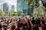 A large crowd of people gathered in an urban park area, holding signs and raising their fists in a gesture of solidarity. The background features tall glass buildings and green trees, suggesting a city setting. The diverse group exhibits a sense of unity and purpose.