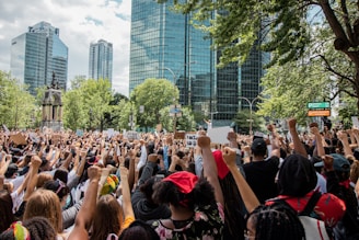 A large crowd of people gathered in an urban park area, holding signs and raising their fists in a gesture of solidarity. The background features tall glass buildings and green trees, suggesting a city setting. The diverse group exhibits a sense of unity and purpose.