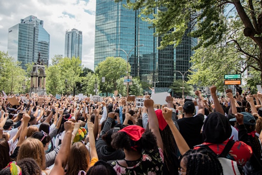 A large crowd of people gathered in an urban park area, holding signs and raising their fists in a gesture of solidarity. The background features tall glass buildings and green trees, suggesting a city setting. The diverse group exhibits a sense of unity and purpose.