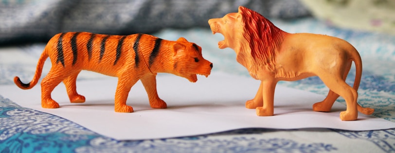 Two toy animals, a tiger and a lion, stand facing each other on a piece of paper. The tiger has an orange and black striped pattern, while the lion has a yellow body and a dark mane. The background features a blurred, patterned fabric.