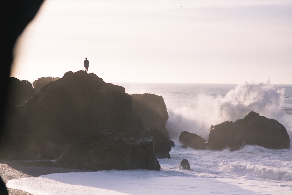 silhouette di persona in piedi sulla formazione rocciosa vicino al mare durante il giorno