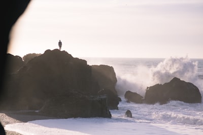 silhouette of person standing on rock formation near sea during daytime vast teams background