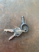 Two metallic keys attached to a black plastic key ring are resting on a textured, dark surface. The keys have numbers and letters engraved on them.