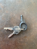 Two metallic keys attached to a black plastic key ring are resting on a textured, dark surface. The keys have numbers and letters engraved on them.