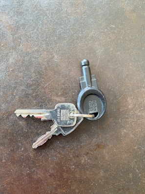 Two metallic keys attached to a black plastic key ring are resting on a textured, dark surface. The keys have numbers and letters engraved on them.