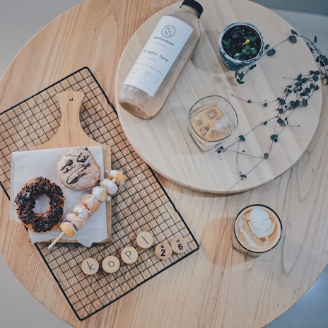 A wooden table holds a variety of breakfast items including a bottle of iced coffee, a glass of iced beverage, and a cup of cappuccino topped with foam. A metal grid rests on a wooden board displaying a powdered skewer, a chocolate-covered donut, and a bun with chocolate drizzle. Small wooden disks with letters spell ‘KOPI 26’. A small potted plant and some decorative twigs add a touch of greenery.