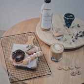 An arrangement of coffee items and pastries on a wooden table. There is a bottle of iced coffee with a label, a glass partially filled with a beverage, and a cup of hot coffee with foam art. A wire rack holds a chocolate-glazed donut, an amusingly decorated cookie with a face, and a skewer of dough balls dusted with powdered sugar. Wooden discs with letters spell out 'K O P I' and the numbers '2' and '6'. A small potted plant and a dried branch add a touch of nature to the setting.