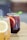 Two glasses of refreshing beverages are placed on a wooden surface. The glass in the foreground contains a light-colored drink with a lemon slice, while the glass behind it holds a dark red drink garnished with a slice of orange and a sprig of mint. Both glasses have black straws.