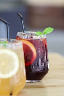 Two glasses of refreshing beverages are placed on a wooden surface. The glass in the foreground contains a light-colored drink with a lemon slice, while the glass behind it holds a dark red drink garnished with a slice of orange and a sprig of mint. Both glasses have black straws.