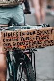A close-up shot of a bicycle with a cardboard sign attached to the back. The sign has a handwritten message that reads: 'Holes in our spirit cause in tears + fears one-sided stories, for years. Take the power.' The legs of a person in shorts are visible in the background.