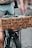 A close-up shot of a bicycle with a cardboard sign attached to the back. The sign has a handwritten message that reads: 'Holes in our spirit cause in tears + fears one-sided stories, for years. Take the power.' The legs of a person in shorts are visible in the background.