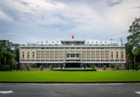 A large, rectangular government building surrounded by a lush green lawn. The facade features vertical lines and ornamental details, with a prominent red and yellow flag flying on the rooftop. Flanking the building are a series of red flags on poles set against a backdrop of trees. The sky is cloudy, casting a soft light over the scene.