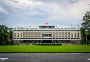 A large, rectangular government building surrounded by a lush green lawn. The facade features vertical lines and ornamental details, with a prominent red and yellow flag flying on the rooftop. Flanking the building are a series of red flags on poles set against a backdrop of trees. The sky is cloudy, casting a soft light over the scene.