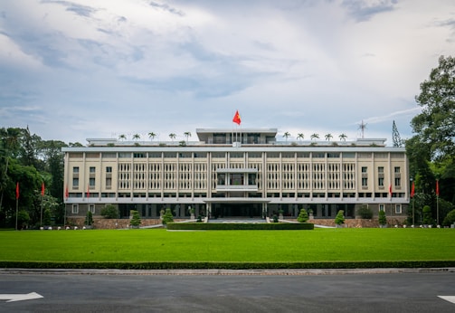 A large, rectangular government building surrounded by a lush green lawn. The facade features vertical lines and ornamental details, with a prominent red and yellow flag flying on the rooftop. Flanking the building are a series of red flags on poles set against a backdrop of trees. The sky is cloudy, casting a soft light over the scene.