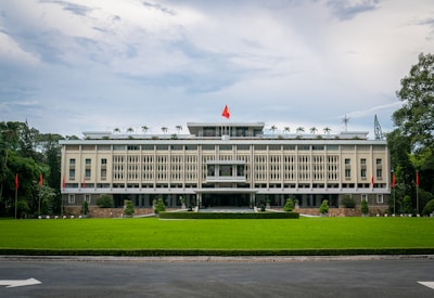 A large, rectangular government building surrounded by a lush green lawn. The facade features vertical lines and ornamental details, with a prominent red and yellow flag flying on the rooftop. Flanking the building are a series of red flags on poles set against a backdrop of trees. The sky is cloudy, casting a soft light over the scene.
