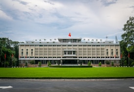 A large, rectangular government building surrounded by a lush green lawn. The facade features vertical lines and ornamental details, with a prominent red and yellow flag flying on the rooftop. Flanking the building are a series of red flags on poles set against a backdrop of trees. The sky is cloudy, casting a soft light over the scene.