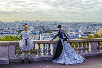 Two individuals dressed in bold, avant-garde fashion pose on a stone balustrade overlooking a panoramic cityscape. The outfits feature striking black and white stripes with elements of modern design, set against a background of a sprawling urban landscape under a cloudy sky.