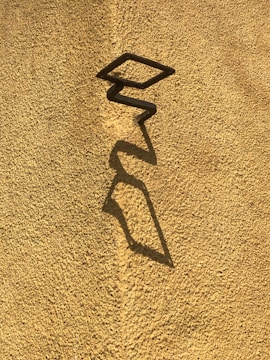 A textured yellow wall features a zigzagging shadow created by a protruding angular metal structure, casting an elongated and distorted shape on the surface.