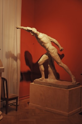 A classical marble statue of a male figure in a dynamic pose, with one arm extended forward as if reaching out. The statue is mounted on a rectangular pedestal against a reddish-brown wall, with soft lighting casting dramatic shadows. A wooden chair and a draped curtain are partially visible to the side, adding to the setting's classical and museum-like ambiance.