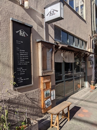 A quaint storefront features a vertical black signboard with white text, flanked by a small wooden bench and planters. The exterior has a beige facade and a box light with Japanese characters hangs above. Displayed near the entrance are menus and photographs inside a wooden frame.