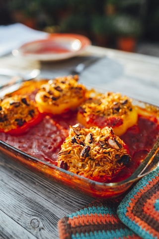 A glass baking dish containing baked stuffed bell peppers with rice and beans on a wooden table. The stuffing appears golden and slightly crispy, surrounded by a rich, red tomato sauce. The setting suggests a rustic outdoor meal, with a knitted pot holder nearby and warm sunlight creating a cozy atmosphere.