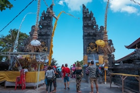 A traditional Balinese gate adorned with intricate stone carvings and colorful ceremonial decorations, including umbrellas and fabrics. Several people are gathered near the gate, some taking photos and others walking through.