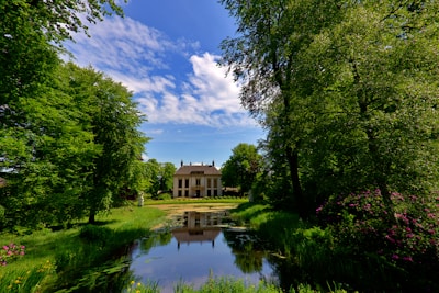 A symmetrical scene featuring a large, elegant house with classical architecture, framed by lush green trees on either side. A tranquil pond in the foreground reflects the building and the vivid blue sky with scattered clouds.
