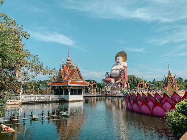 A serene temple complex featuring a large, smiling Buddha statue with an ornate halo. The statue overlooks a still, reflective body of water adorned with lotus-like structures. A traditional temple building with an intricate roof design is prominently situated by the water's edge, and lush greenery surrounds the area.