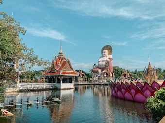 A serene temple complex featuring a large, smiling Buddha statue with an ornate halo. The statue overlooks a still, reflective body of water adorned with lotus-like structures. A traditional temple building with an intricate roof design is prominently situated by the water's edge, and lush greenery surrounds the area.