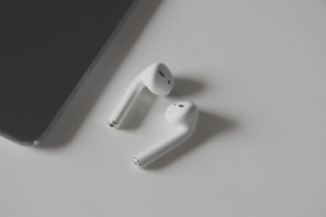 A pair of white wireless earbuds placed on a light grey surface with a black tablet partially visible on the left side.
