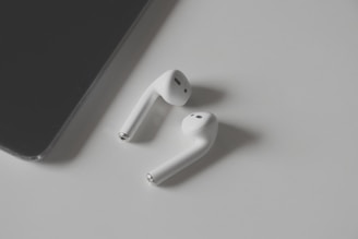 A pair of white wireless earbuds placed on a light grey surface with a black tablet partially visible on the left side.