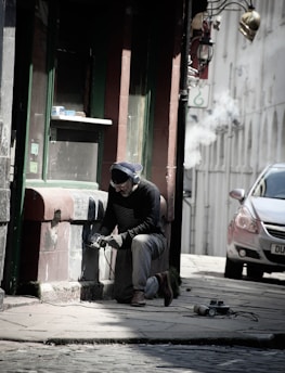 A person kneels on the ground while using a power tool on a building's exterior wall, wearing protective goggles, gloves, and headphones. The scene is set in a narrow street with cobblestone pavement, next to an old building with a green and brown facade. A car is parked nearby, and there are signs of renovation or maintenance work being carried out.