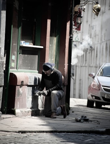 A person kneels on the ground while using a power tool on a building's exterior wall, wearing protective goggles, gloves, and headphones. The scene is set in a narrow street with cobblestone pavement, next to an old building with a green and brown facade. A car is parked nearby, and there are signs of renovation or maintenance work being carried out.