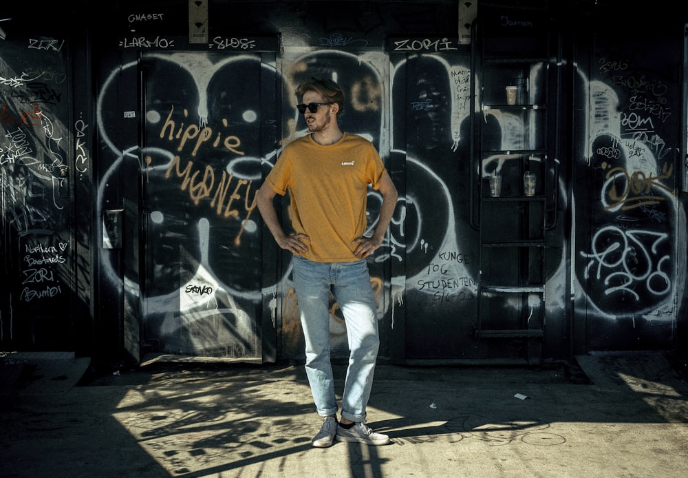 man in orange crew neck t-shirt and blue denim jeans standing beside black wall